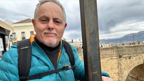 Simon Riggs dressed in a turquoise jacket, with an old bridge in the background