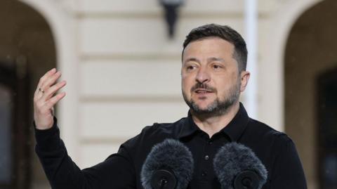 Volodymyr Zelensky speaking at a lectern with his hand raised