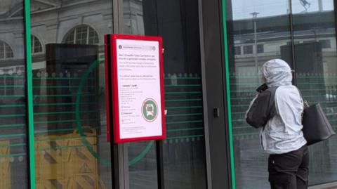 A person wearing a raincoat looks at a sign in front of the Cardiff Bus Interchange. The sign has a red frame. Behind it are closed glass doors and a sign that says Welcome.