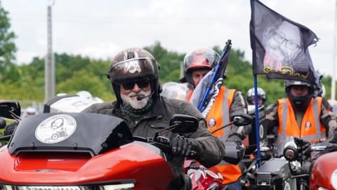 Si King wears a crash helmet as he sits on his motorbike ahead of a procession of other bikers taking part in the first Dave Day. King has a grey beard and is wearing a dark leather jacket. One of the bikes in the background has a flag showing Dave Myers' face.