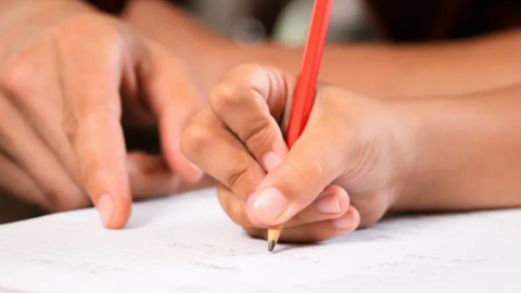 A child's hand holding a red pencil while an adult points at a white paper.
