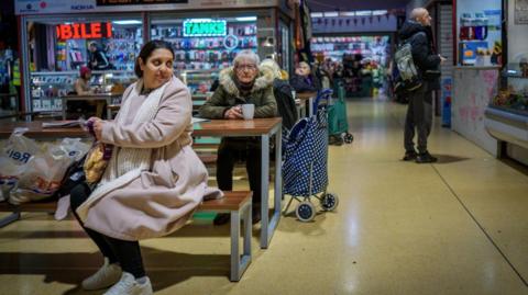People do their shopping in Gorton Market on January 28, 2026 in Gorton, United Kingdom. A woman can be seen sat on a bench with her shopping while an older woman sits with a cup of tea behind her. Others can be seen looking at stalls. 
