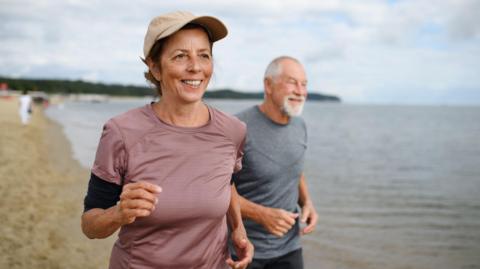 Active senior couple runners jogging outdoors on sandy beach by sea in early morning.