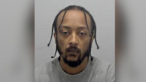 A mugshot of a man wearing grey with braised hair and a thick beard