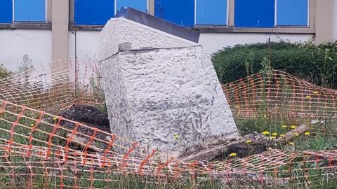 A tipped-over war memorial surrounded by orange plastic barrier fencing which is loose and untidy. There is soil uprooted at the bottom of the concrete base and there appears to be some chipped damage on the stone plinth.