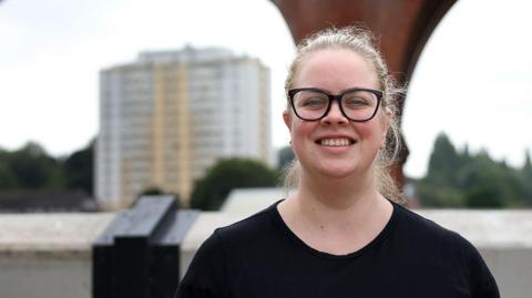 Claire, who is wearing black glasses and a black t-shirt, smiles while standing some considerable distance in front of Stockport's viaduct. Trees and a large block of flats can also be seen behind her. 
