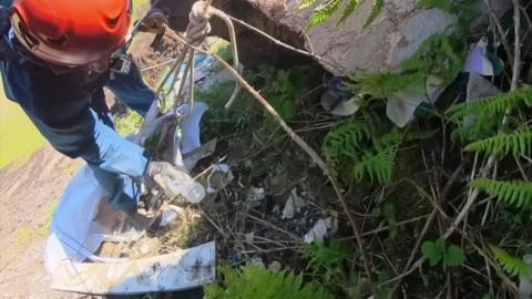 A man in a blue jumpsuit and wearing a red helmet is strapped to a harness on the side of the mountain, and can be seen picking up rubbish as it is strewn across the mountainside.
