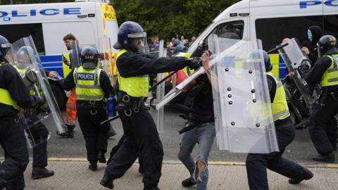 Police officers with protesters as trouble flares during an anti-immigration demonstration. There are two police vans with police in neon vests and helmets in front of them. Two look like they are holding back a person with ripped jeans. A crowd of people stand in the background below green leaves.