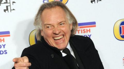 Rik Mayall in front of a British Comedy Awards backdrop, laughing and pointing