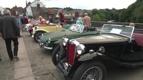 A row of classic cars line the pavement. They are varying makes and colours and all open top. Members of the public mill around looking at them.