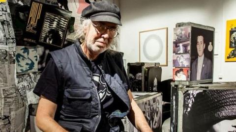 A man wearing a black cap, a black gilet and a black top is looking into the camera with a serious expression and wearing large clear-rimmed glasses. Posters and old photographs are all around him.