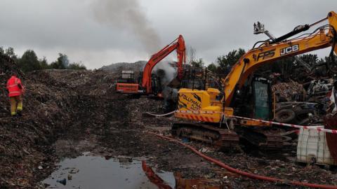 Excavators are parked in the middle of a large waste site. A man in a high vis jacket can be seen walking through it.
