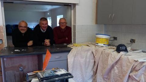 Three male venue trustees smiling and leaning on the worktop window of the new kitchen with sheets, paint and working tools in the kitchen area