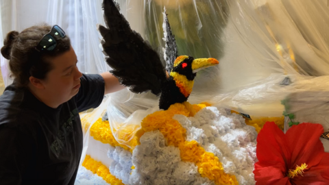 A woman adjusts the positioning of some flowers on a float. The piece is shaped like a black bird with large wings. There are white, yellow and red flowers attached to the float. The woman is wearing a black tshirt, with dark hair and sunglasses on her head. 