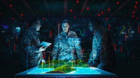 Three people, a woman and two men, all dressed in Army fatigues, stand around a table.  The table is showing a computer simulated landscape.