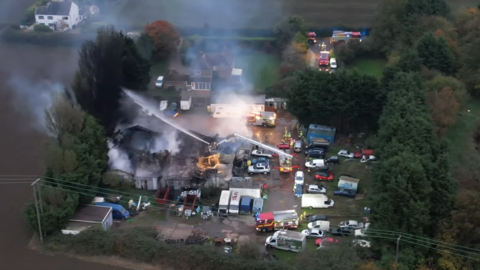 Drone footage shows the fire and crews at the scene from above. There is a large building, which looks to have been mostly charred and burnt, being sprayed with water from fire hoses. The roof of the building has collapsed and there are several plumes of smoke coming from where the roof once was. The are lots of cars around the business - unaffected by the fire - and it looks to be in a rural setting.