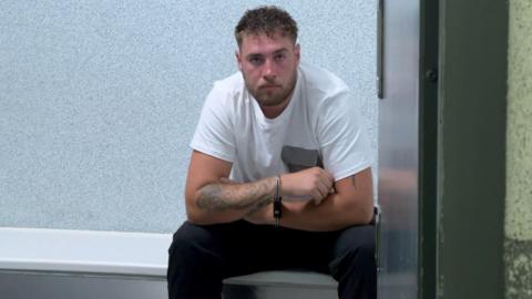 Lewis Carver, wearing a white T-shirt and dark trousers, sits looking glum in handcuffs in a police cell. He has dark hair, a beard and tattoos on his right arm.