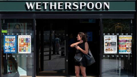 A woman dressed in a black vest and shorts stands outside a Wetherspoons holding her phone.