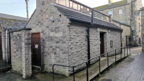 The picture shows an oblong-shaped public toilet built from stone coloured blocks. The small building has a tiled, pitched roof with a row of windows along the top. A warning notice has been stuck to each of the block's two visible doors which say the toilets have closed.