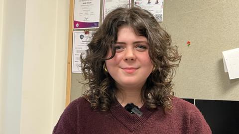 A young woman with curly brown hair and wearing burgundy jumper, smiling at the camera in a classroom with posters on the wall 