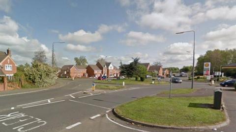 A road with houses along the left hand side and a garage up and on the right. There is a roundabout with a silver car going round it and grass verges with trees in the background. 