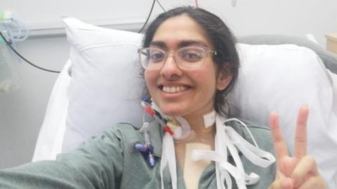 Mehreen Ahmed in a hospital bed with tubes around her neck following her heart transplant. She is wearing glasses and a green gown and is smiling into the camera with her fingers raised in a V for victory sign.