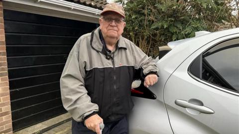 A man leaning against a grey car while holding a walking stick. He is wearing a black and grey coat and a hat.