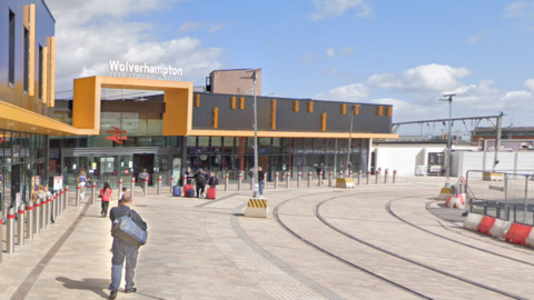 Passengers make their way to the train station in Wolverhampton. It has a yellow and black entrance with an old British Rail sign above the entrance and tram lines running past the front a few feet away.