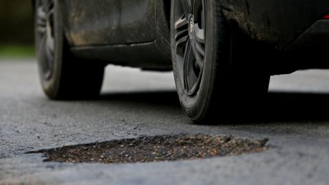 The wheels of a car, viewed from a low angle, travel past a pothole in the road. The car is black, and has mud splattered on its sides.