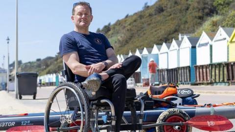 Will sat in his wheelchair with a paddleboard and equipment behind him on the beach