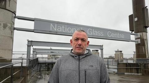 Councillor Phil Tye standing in front of the entrance to the National Glass Centre. He is wearing a grey hoodie. The centre's sign hangs above the entrance supported by two large towers.