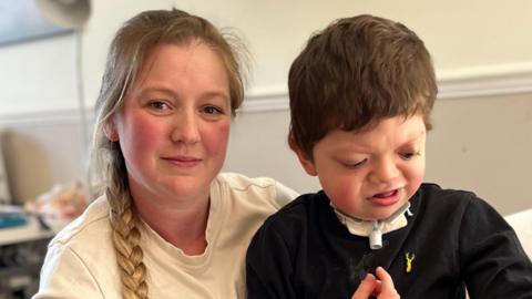 A woman with long plaited hair is sitting on her sofa with her disabled son. She is looking intently into the camera with her 10 year old son on her knee. He has a facial deformity and a tube is attached to his throat. He is looking away from the camera. 