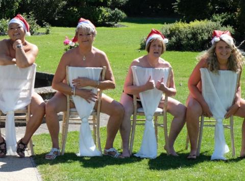 A group of woman, who are naked, sat in chairs that are the wrong way around and covering most of their bodies.