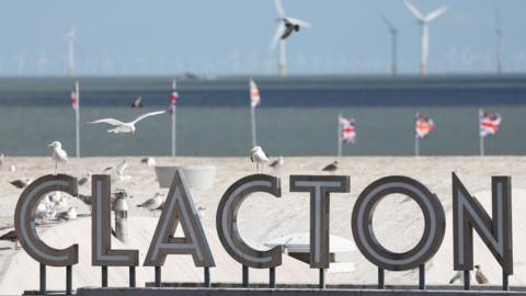 Seagulls perch on and fly above a sign that reads "CLACTON". In the background, six union jacks are flying. Beyond that is the sea and a number of wind turbines.