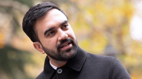 Zohran Mamdani in a grey coat mid-speech as he stands outdoor, yellow foliage in the blurred background
