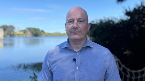 Jon Holt is a bald man looking at the camera with a slight smile. He is wear a blue shirt. You can see a body of water behind him.