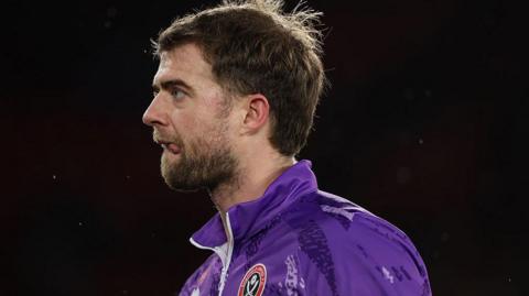 Sheffield United striker Patrick Bamford, bearded, and with his tongue sticking out of the side of his mouth, wearing purple Shewffield United tracksuit top and staring forward
