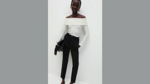 M&S model poses in white top and black trousers and leans against a white wall