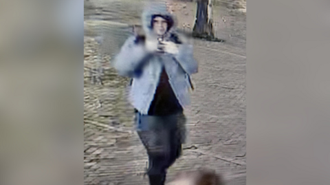 The image shows a blurry CCTV image of a man in a grey jacket and black bottoms. He is walking and the trunk of a tree can be seen in the background.