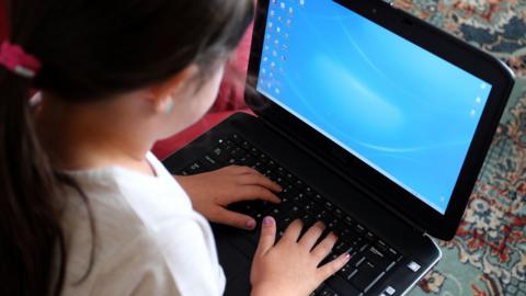 A small girl with a pony tail hairstyle and pink nail varnish uses a laptop.