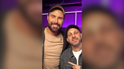 Rylan with Dynamo in the purple-lit radio studio, both wearing caps, Dynamo pointing at Rylan as they pose for a photo.