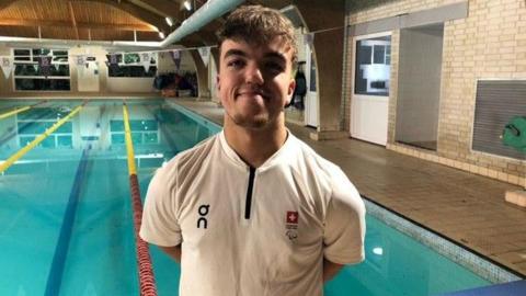 Leo McCrea posing for the camera in front of a swimming pool. He has short dark hair and is wearing a white t-shirt with a small Swiss flag logo on the chest.