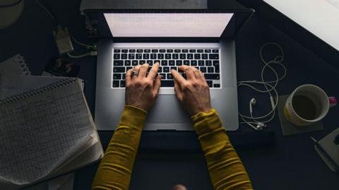 A woman is sitting at a desk and is typing on the keyboard of a laptop. There is a set of small headphones and a coffee cup next to the computer. A pad of paper with a grid drawn on is also on the desk.