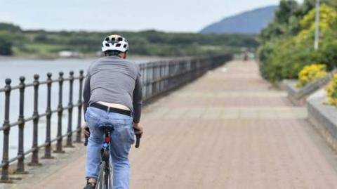 A man riding a bicycle along a path by a river. There is a metal railing separating him from the river. Trees and shrubbery line the path on the other side.