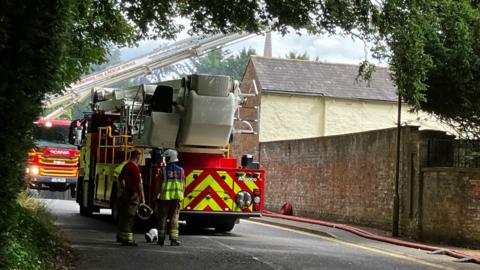 The back of a fire engine on a lane with two firefighters standing behind it. One man has his helmet off and the other is wearing a helmet and a hi-vis coat. In front of the fire engine is another fire engine with an aerial ladder extending towards the roof of a house.