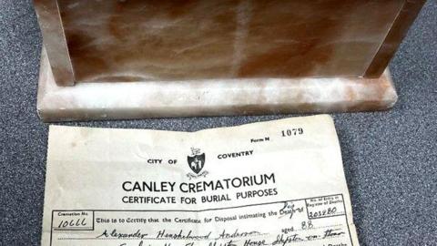 A brown and white marble box and yellowed burial certificate on a grey table top. The name Alexander Anderson and age of 88 can be made out on the certificate.