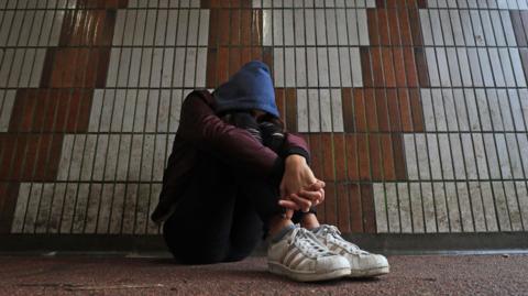 A girl sitting on the ground next to dirty wall tiles in an underpass with her face covered by her knees, with her blue hood pulled over her head. She is wearing white trainers and a purple top, 