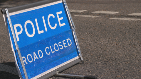 A square blue sign with white writing reads: "Police, road closed". It stands on a grey road surface with white road markings indicating a junction.
