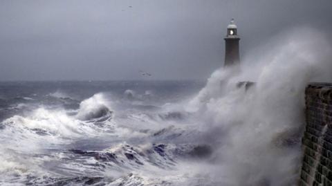 A lighthouse being splashed with large waves, there are seagulls and a cloudy darkening sky