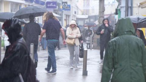 People walking in the rain at Charing Cross in St Helier. Many have umbrellas. Most have raincoats on with their hoods up. Shop signs are visible in the background.